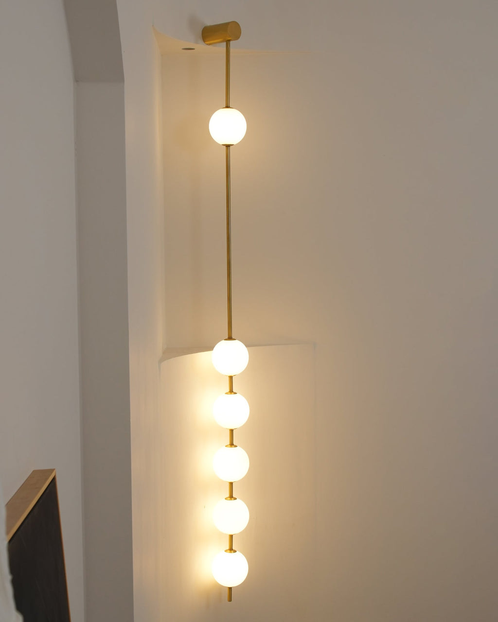 The Clérachic Stacked Globe Wall Sconce in brass, lit, five globes glowing in a stairwell corner beside a handrail.