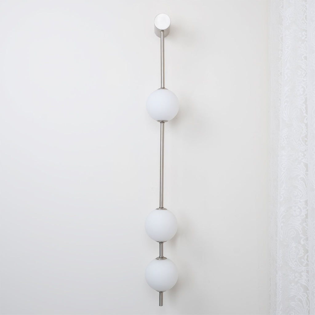 The Clérachic Stacked Globe Wall Sconce in brushed nickel, lit, three globes beside a sheer lace curtain.