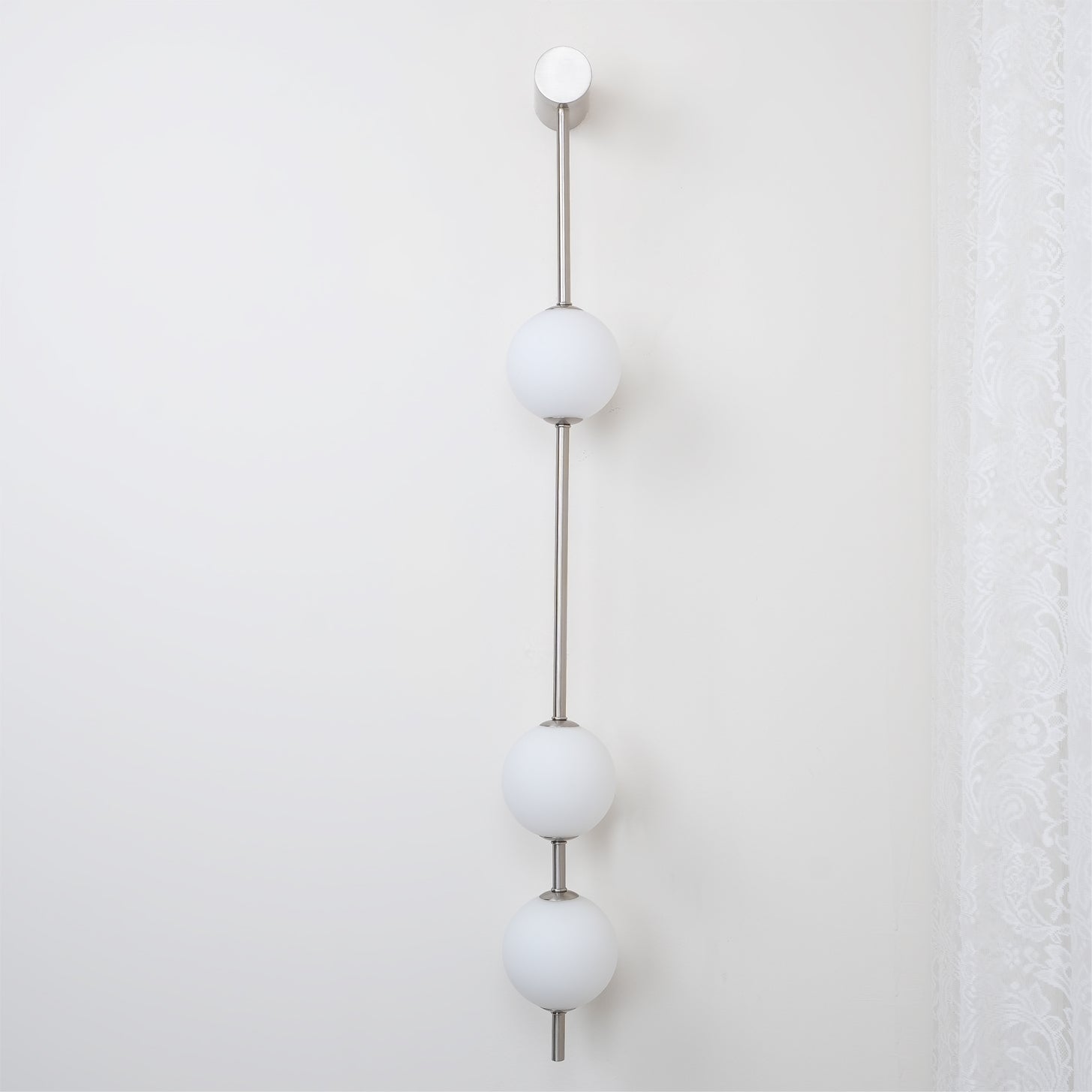 The Clérachic Stacked Globe Wall Sconce in brushed nickel, lit, three globes beside a sheer lace curtain.