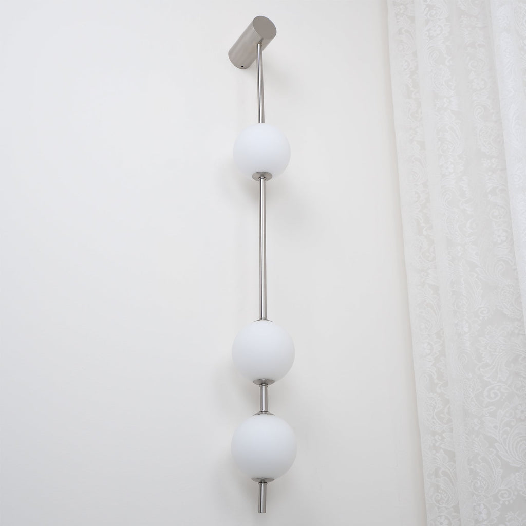 The Clérachic Stacked Globe Wall Sconce in brushed nickel, lit, three globes shown close to a lace curtain.