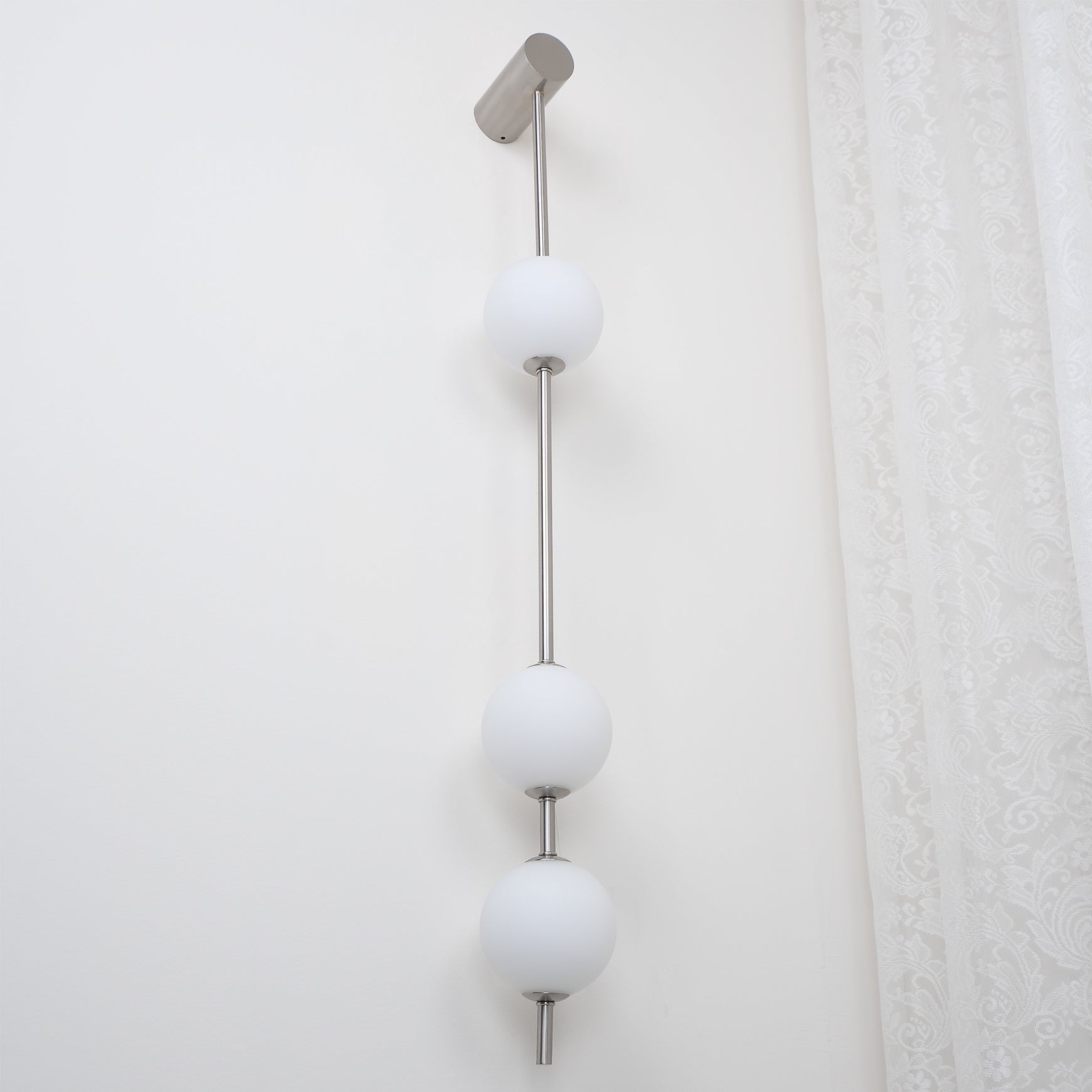 The Clérachic Stacked Globe Wall Sconce in brushed nickel, lit, three globes shown close to a lace curtain.