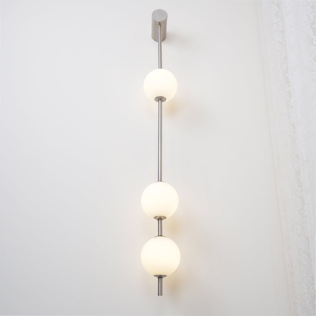 The Clérachic Stacked Globe Wall Sconce in brushed nickel, lit, four globes glowing beside a curtained window.