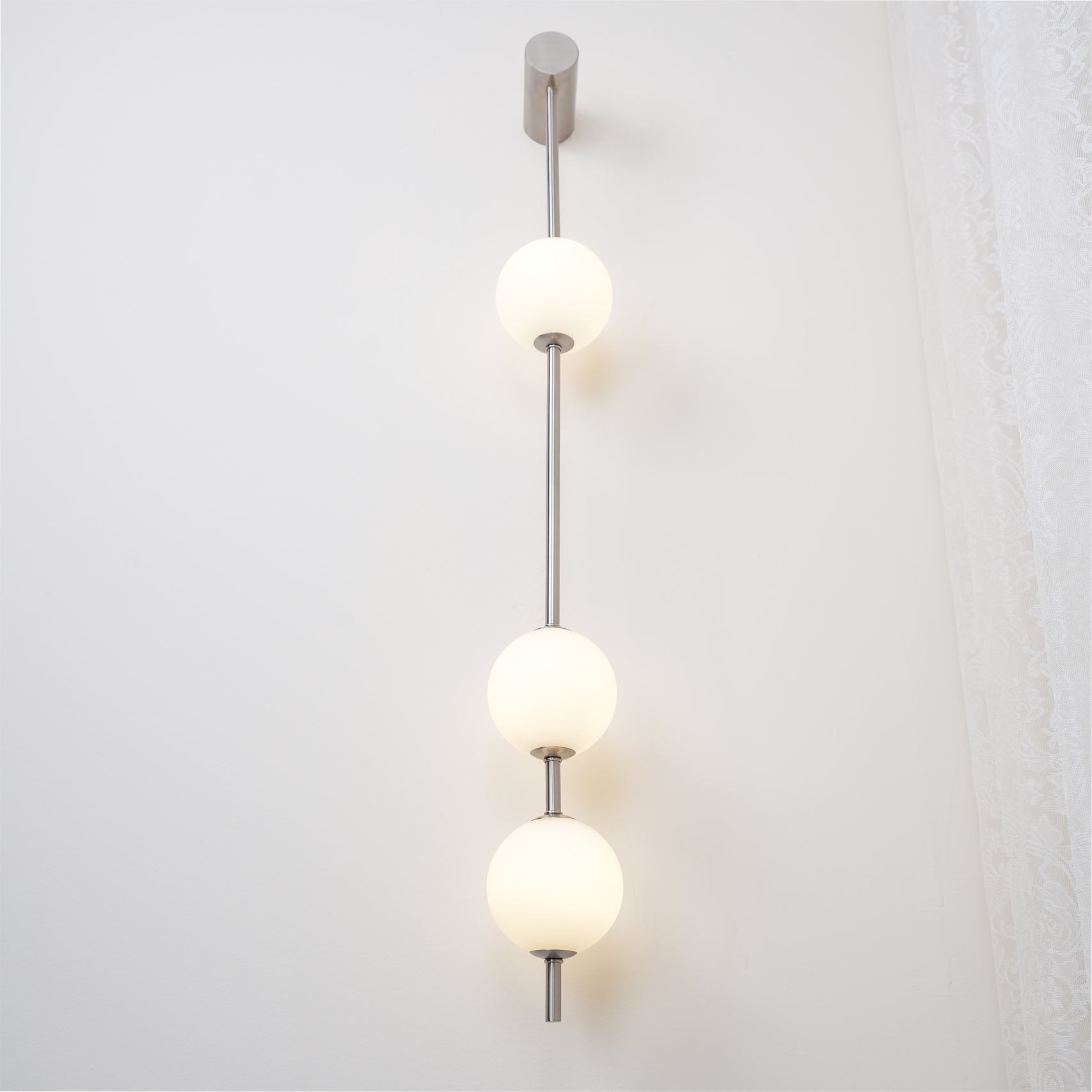 The Clérachic Stacked Globe Wall Sconce in brushed nickel, lit, four globes glowing beside a curtained window.