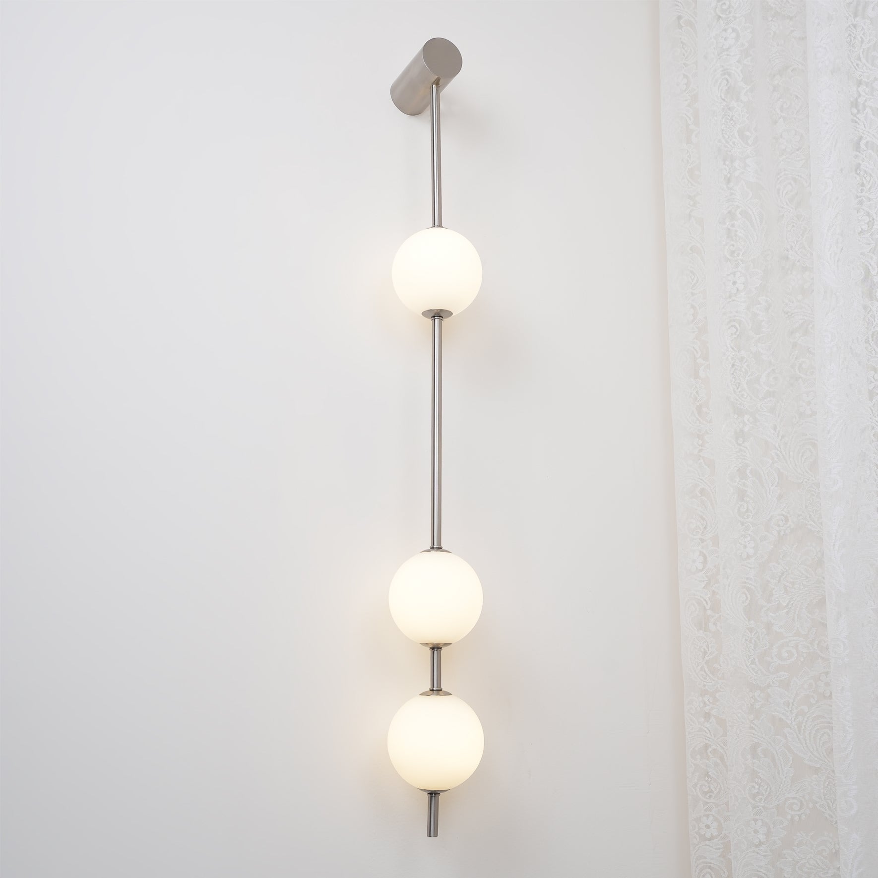 The Clérachic Stacked Globe Wall Sconce in brushed nickel, lit, four globes photographed beside sheer lace.