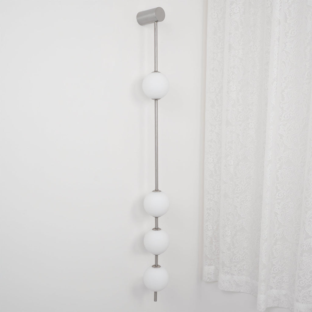The Clérachic Stacked Globe Wall Sconce in brushed nickel, lit, five globes beside a sheer lace curtain.