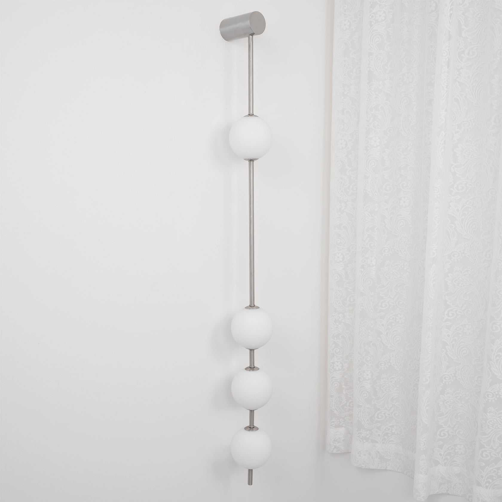 The Clérachic Stacked Globe Wall Sconce in brushed nickel, lit, five globes beside a sheer lace curtain.