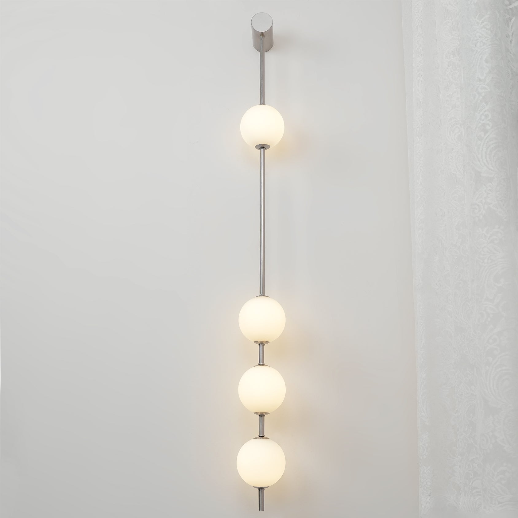 The Clérachic Stacked Globe Wall Sconce in brushed nickel, lit, four globes glowing beside a lace curtain.
