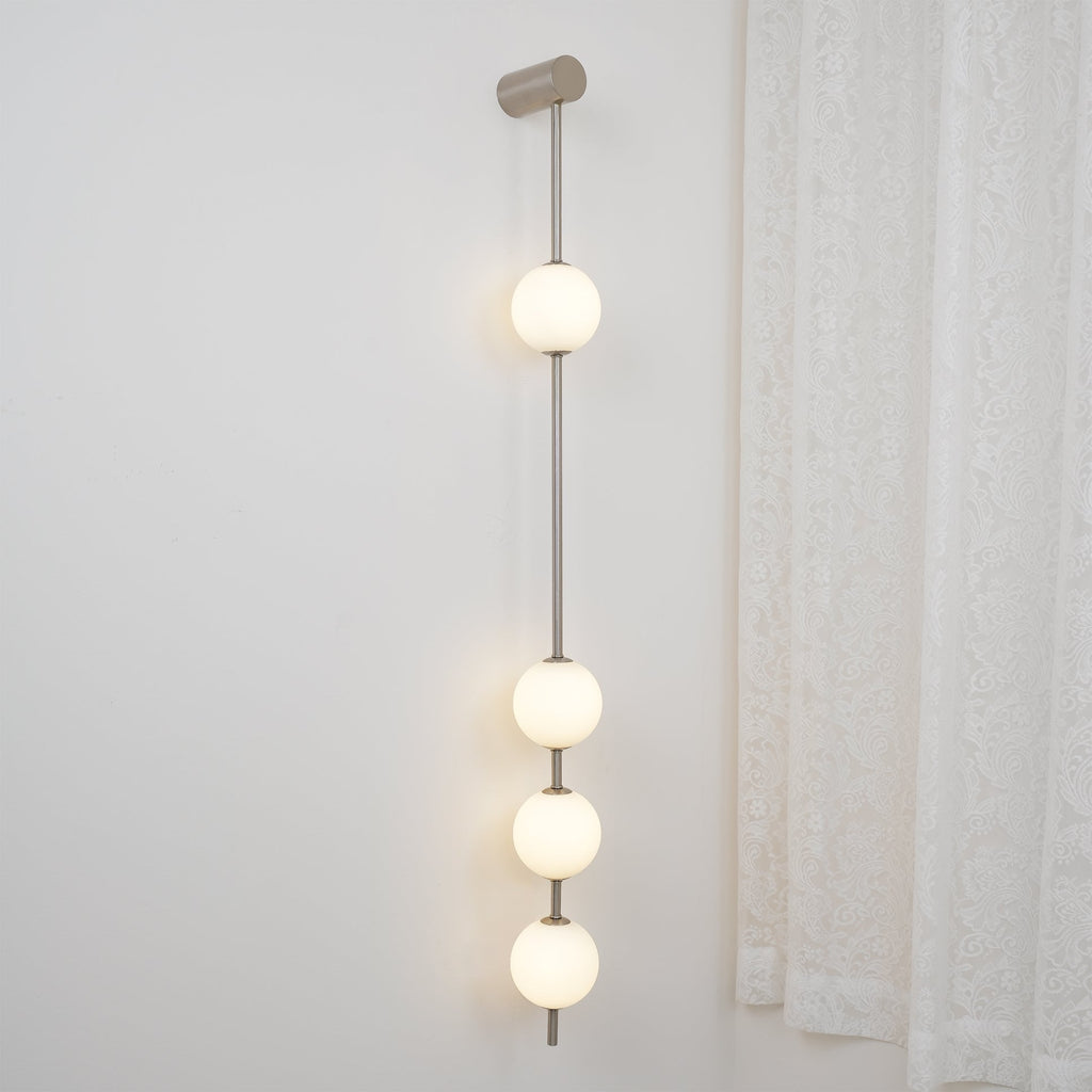 The Clérachic Stacked Globe Wall Sconce in brushed nickel, lit, four globes shown close to a curtained window.