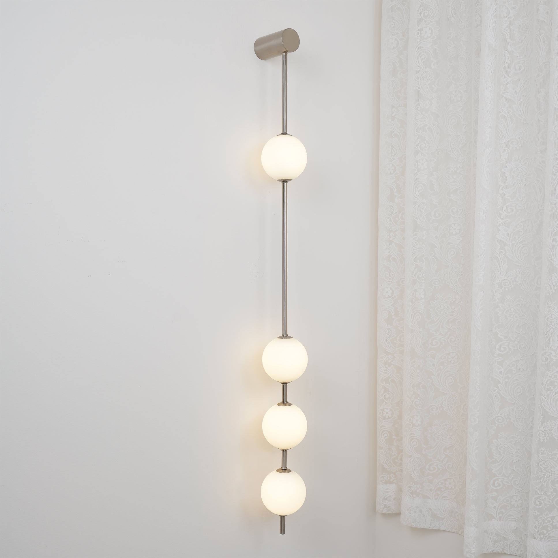 The Clérachic Stacked Globe Wall Sconce in brushed nickel, lit, four globes shown close to a curtained window.