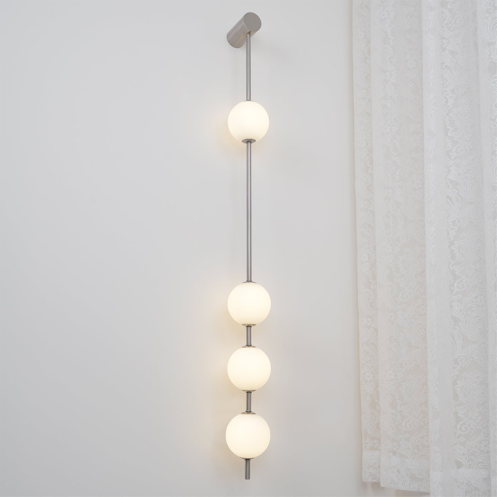 The Clérachic Stacked Globe Wall Sconce in brushed nickel, lit, five globes glowing beside a curtained window.