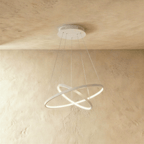 Two white rings cross at an angle in this Clérachic pendant, each edge glowing warm against a plaster wall.