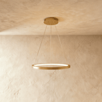 A golden ring pendant by Clérachic glows softly on slender cables below a stone-textured ceiling.