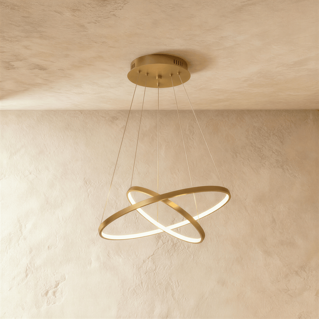 Two golden rings cross at an angle in this Clérachic pendant, each edge glowing warm against a plaster wall.