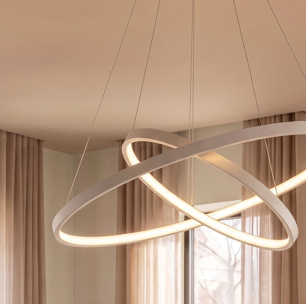 A close-up of Clérachic's white crossed rings glows warm against sheer curtains in a bright living room.