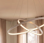 A close-up of Clérachic's white crossed rings glows warm against sheer curtains in a bright living room.