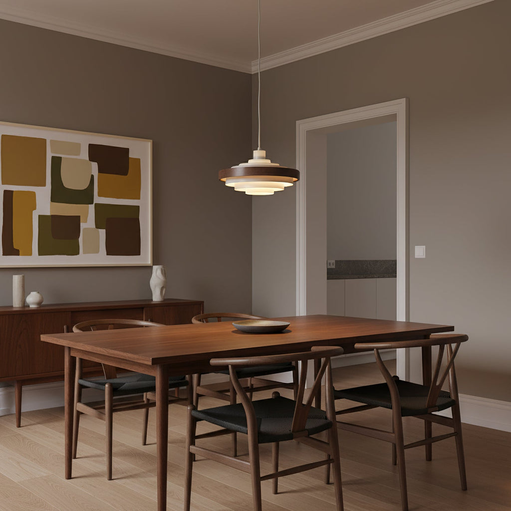 A Clérachic stacked ring pendant hangs over a walnut dining table with wishbone chairs, in a room with an abstract print.