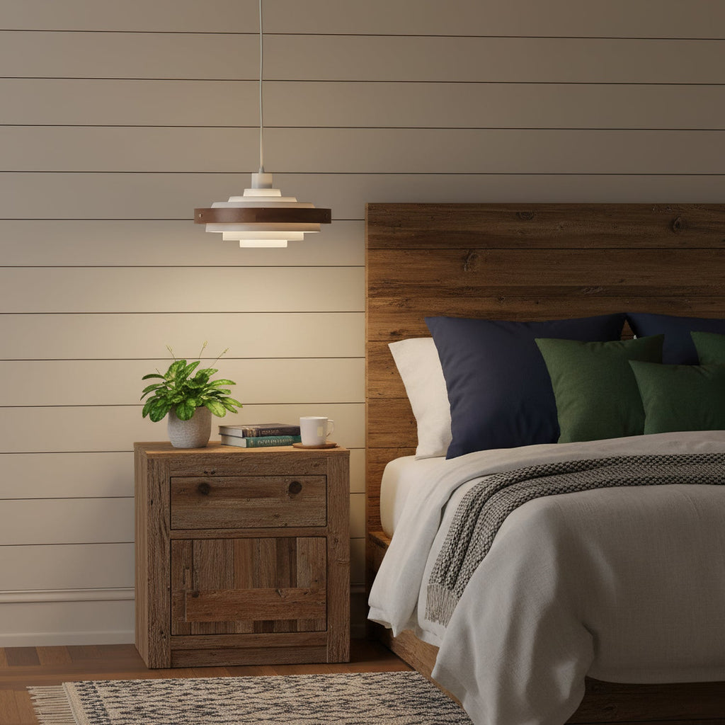 Above a bedside table with a small plant, a Clérachic stacked ring pendant glows beside a bed with a rustic wooden headboard.
