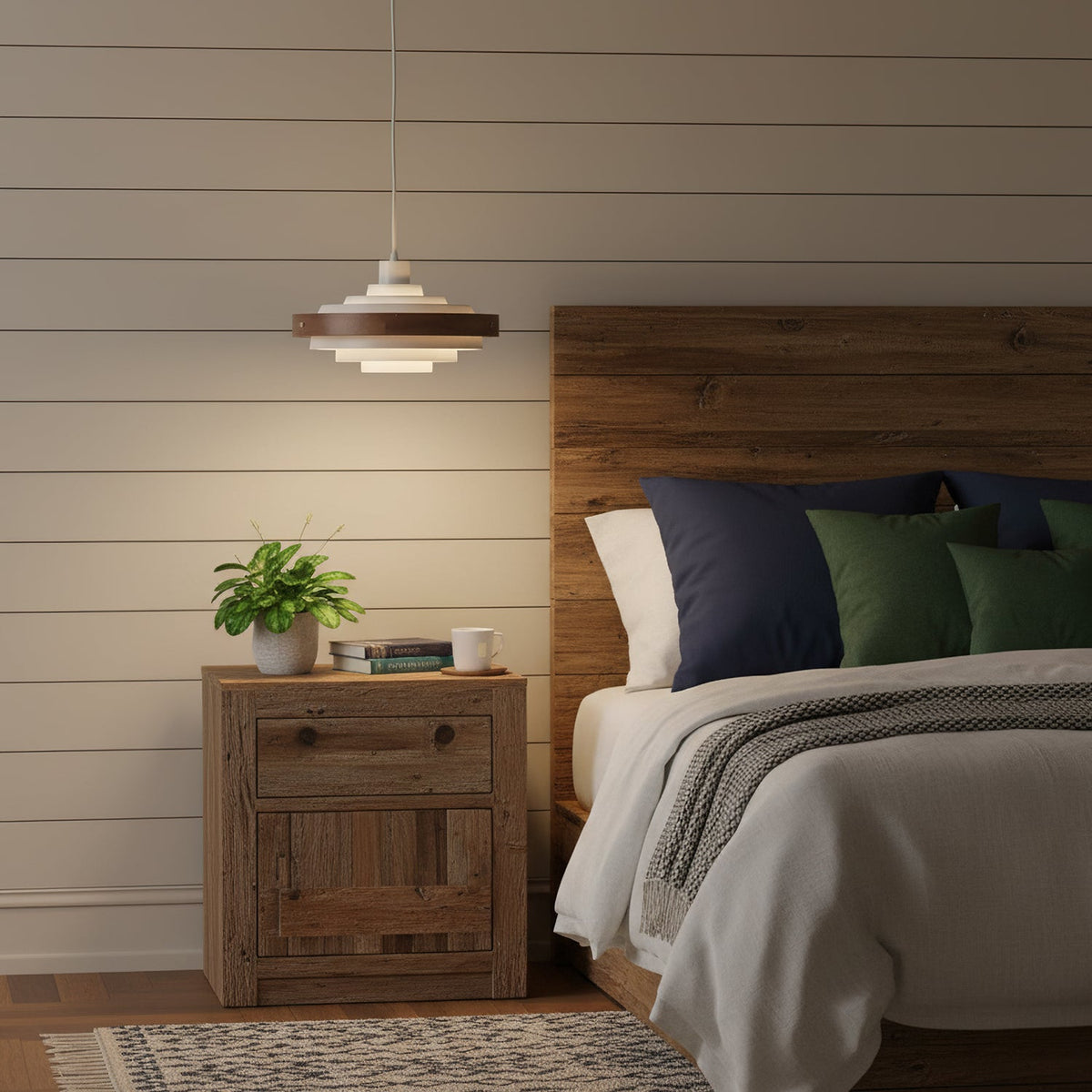 Above a bedside table with a small plant, a Clérachic stacked ring pendant glows beside a bed with a rustic wooden headboard.