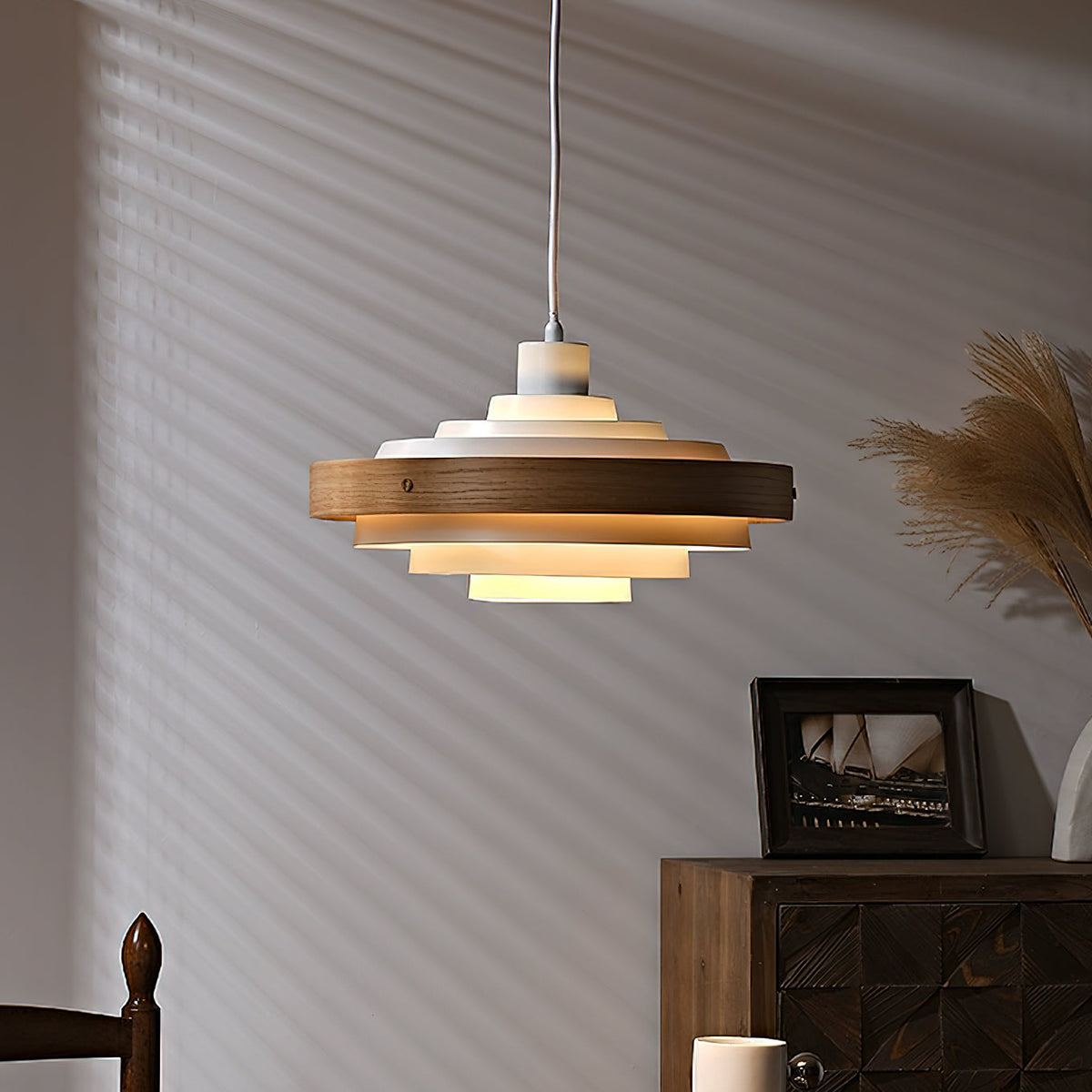 A white and walnut Clérachic stacked ring pendant glows warm in front of a striped shadow wall, with dried pampas grass beside it.