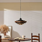 A black and walnut Clérachic stacked ring pendant hangs on a black cord above a small round table with an open book and a mug.