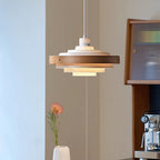 The lit tiers of a Clérachic white and walnut pendant in a room with a glass-fronted wood cabinet and a pink flower in a vase.