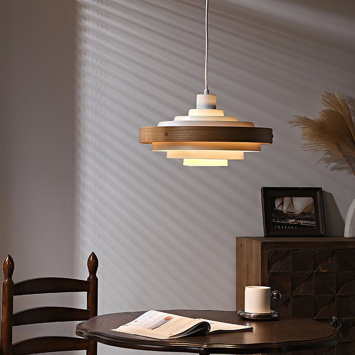 The white and walnut Clérachic pendant glows warm above a dark round table with an open magazine and a cup.