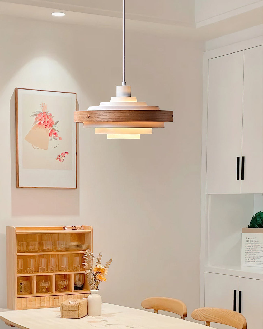 A Clérachic stacked ring pendant in a bright dining corner, with a floral print, white cabinets and a wooden glass shelf.