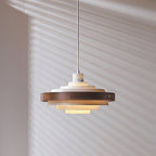 White and walnut Clérachic stacked ring pendant, unlit, hung on a white cord against a wall with striped shadows.