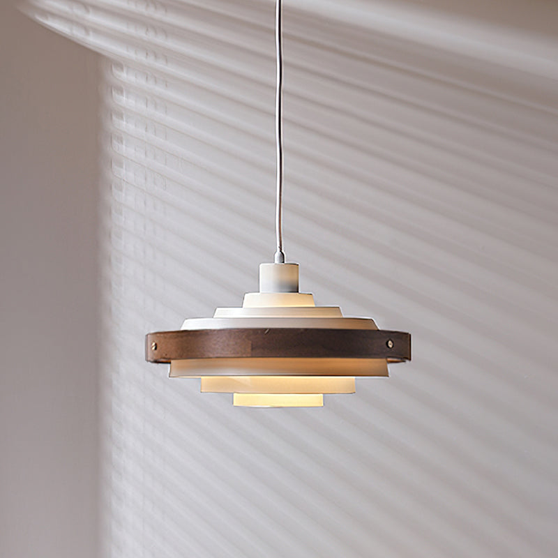 White and walnut Clérachic stacked ring pendant, unlit, hung on a white cord against a wall with striped shadows.
