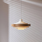 A Clérachic white and oak pendant, lit, on a thin cord in front of a wall with window-blind shadows.