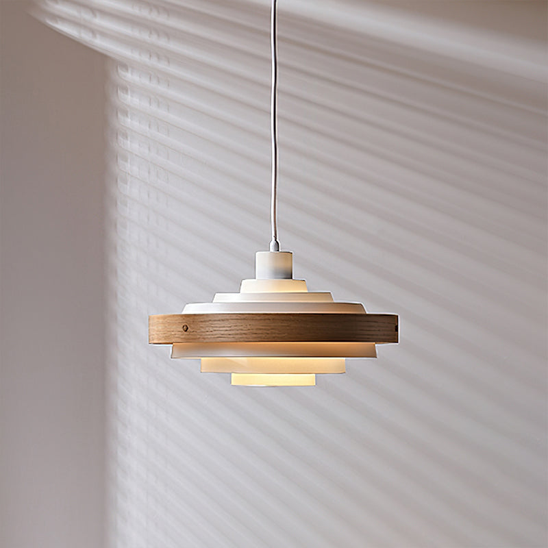 A Clérachic white and oak pendant, lit, on a thin cord in front of a wall with window-blind shadows.