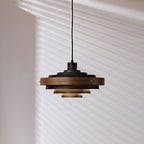 Black and oak Clérachic stacked ring pendant on a black cord, with its lower tiers lit warm, against a wall with blind shadows.