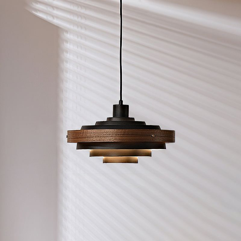 Black and oak Clérachic stacked ring pendant on a black cord, with its lower tiers lit warm, against a wall with blind shadows.