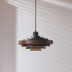Black and walnut Clérachic stacked ring pendant, with the walnut band and brass screws visible, in front of a shadowed wall.