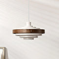 White and walnut Clérachic stacked ring pendant on a white cord, its dark wood band around the top tiers, against a pale wall.