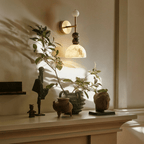 Clérachic wall lamp glowing warm above a mantel styled with terracotta pottery, a carved stone bust and a leafy branch.