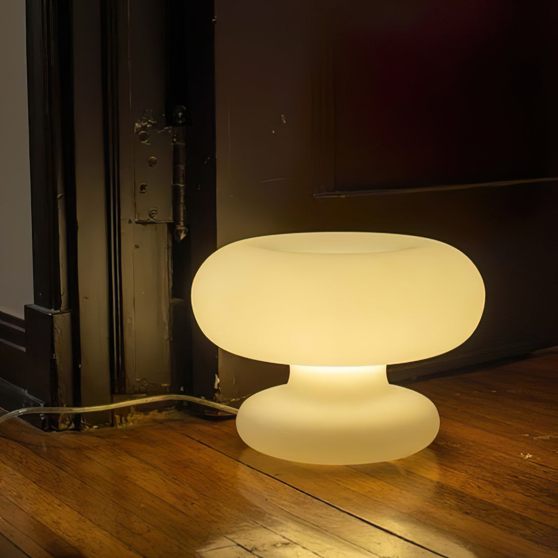 A lit Clérachic donut table lamp on an old wooden floor beside a dark door frame, casting a warm pool of light.