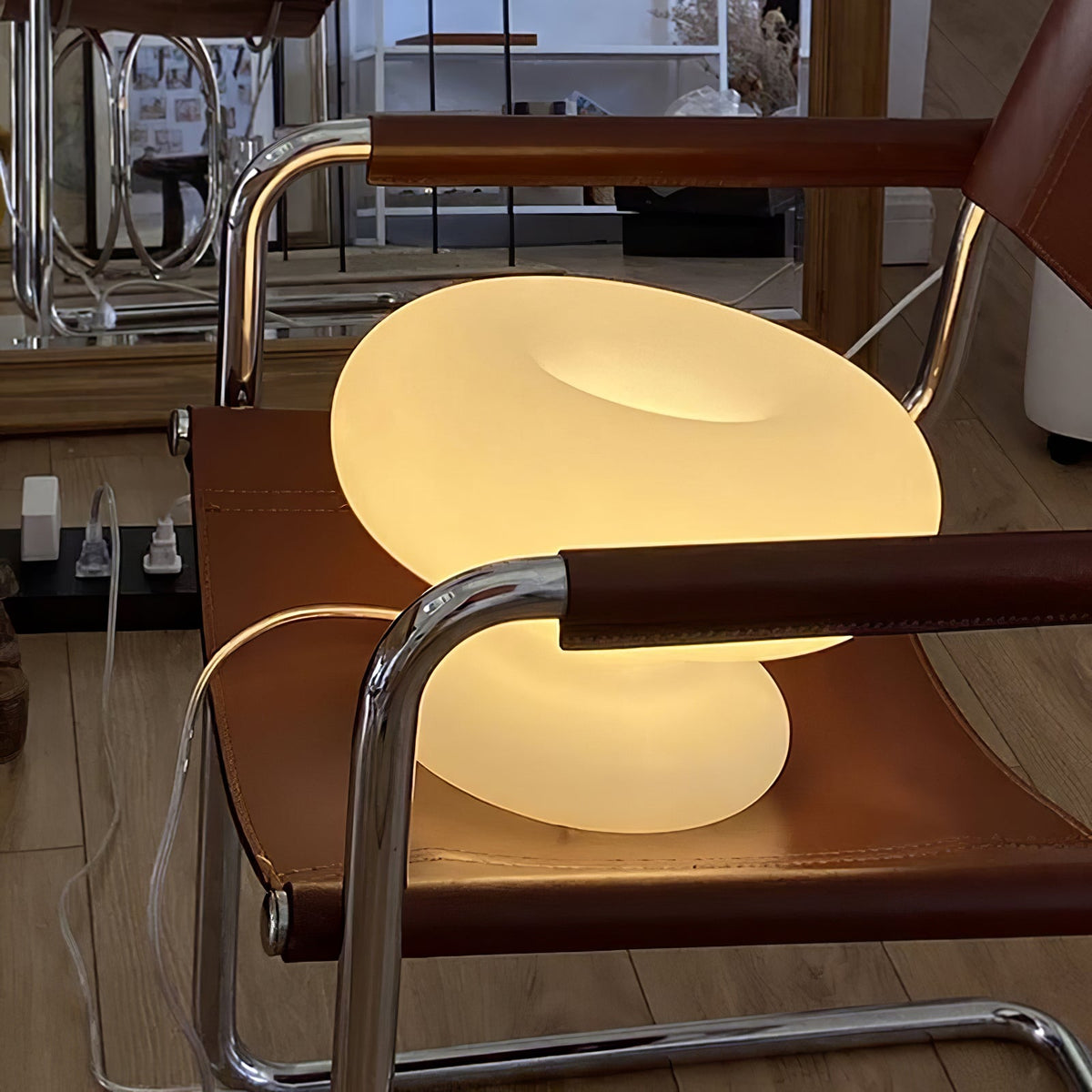 Clérachic donut lamp lit on a tan leather sling chair with chrome arms, its frosted glass glowing a soft warm yellow.