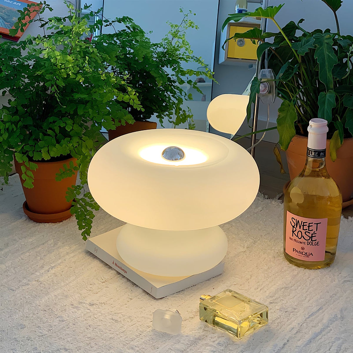 Frosted donut lamp from Clérachic on a white rug among potted ferns, with a bottle of rosé and a small perfume bottle nearby.