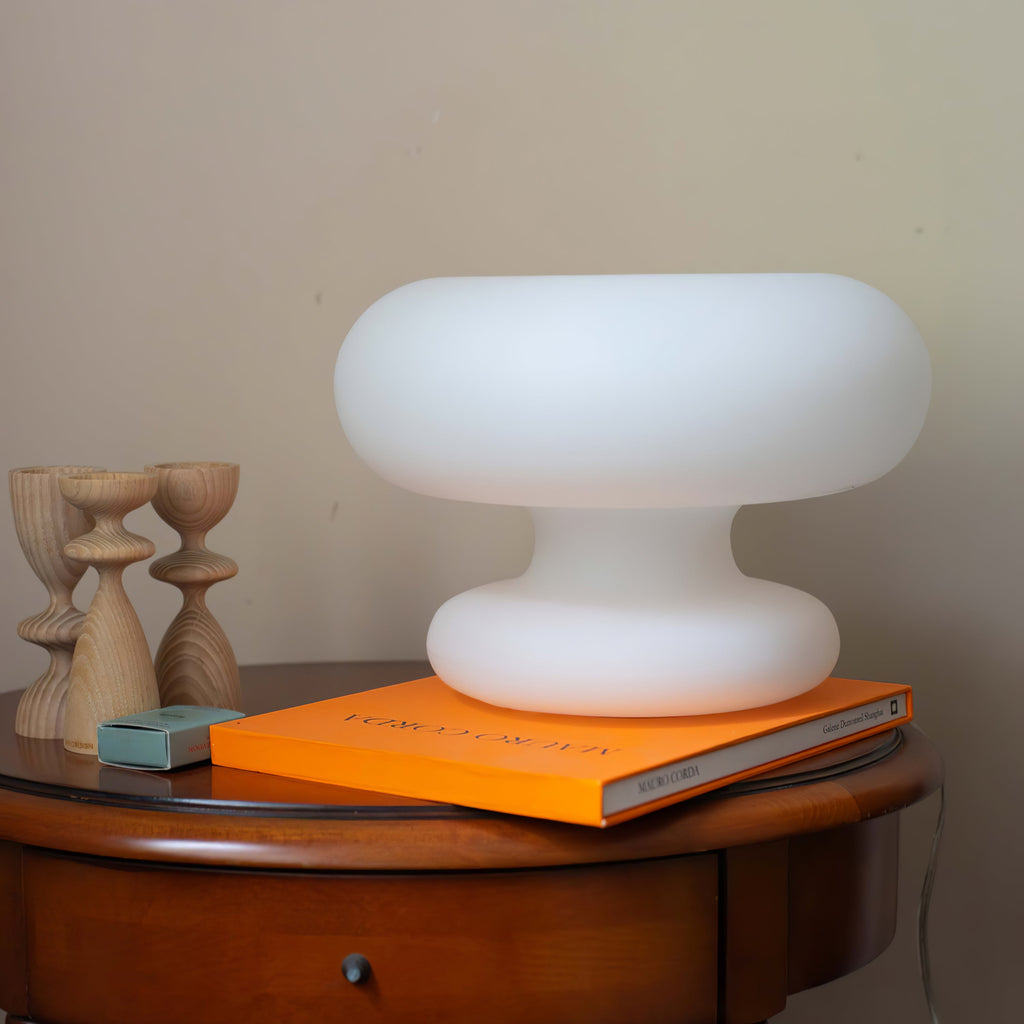 Unlit white Clérachic donut lamp on a round side table, next to an orange art book and two turned wooden candlesticks.