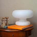 Unlit white Clérachic donut lamp on a round side table, next to an orange art book and two turned wooden candlesticks.