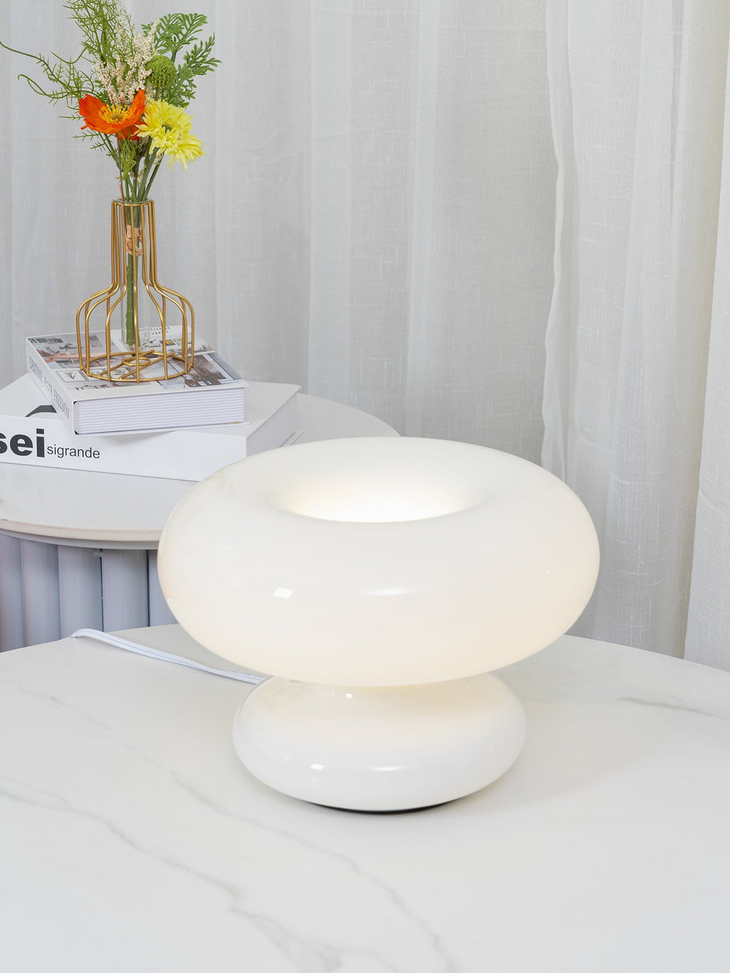 A Clérachic donut lamp glowing softly on a marble-topped table, with a small vase of orange and yellow flowers behind it.