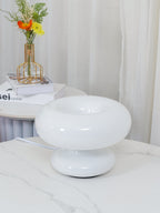 White Clérachic donut lamp switched off on a marble table, with sheer curtains and a gold-framed flower vase behind it.