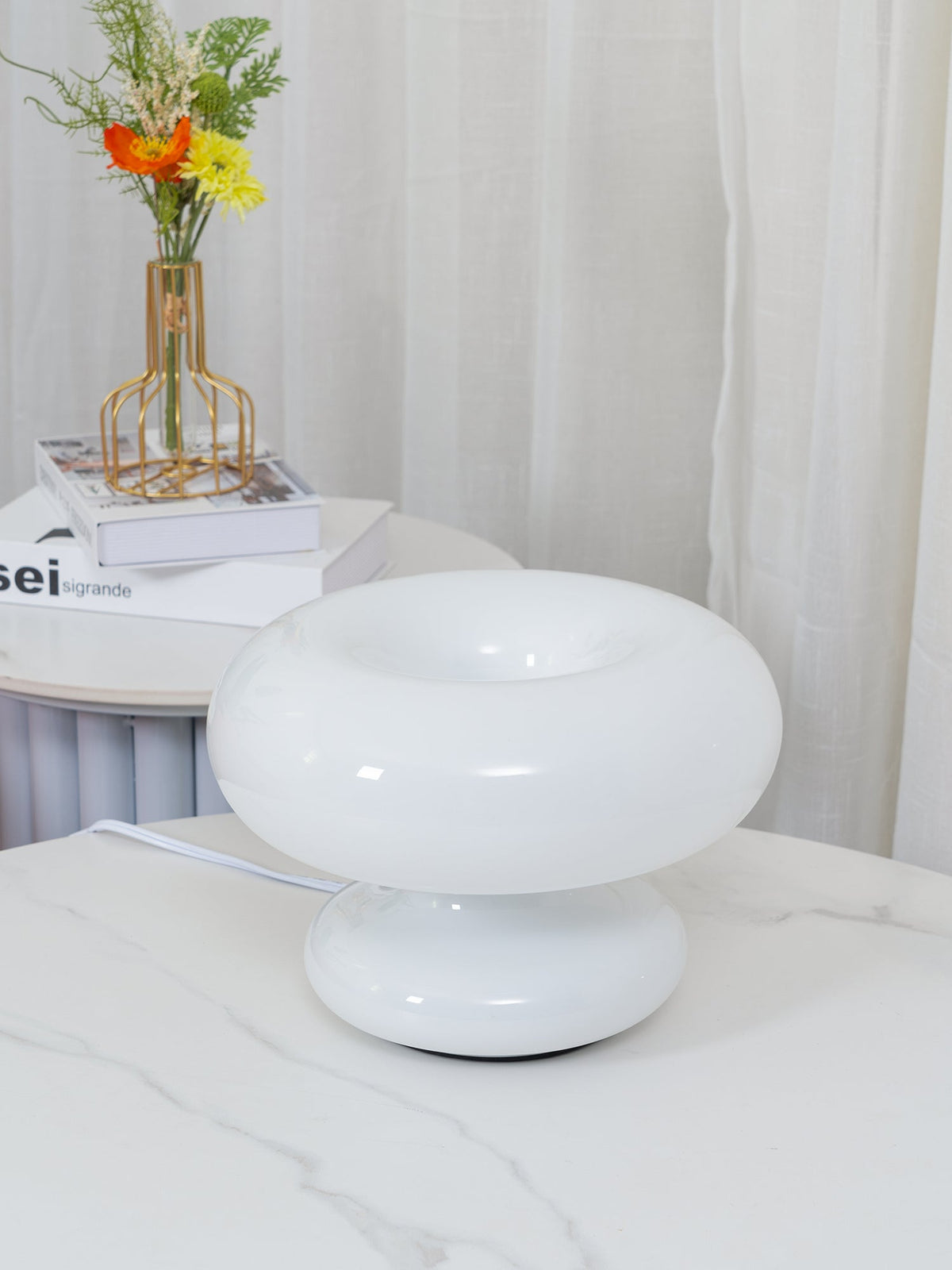 White Clérachic donut lamp switched off on a marble table, with sheer curtains and a gold-framed flower vase behind it.