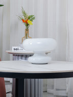 Side view of the stacked white glass Clérachic donut lamp on a marble table, with sheer curtains and flowers in the background.