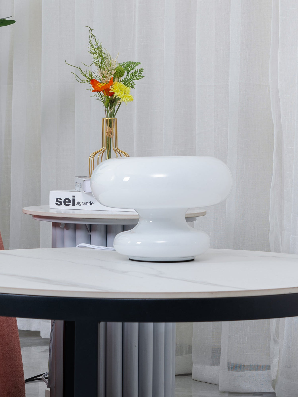 Side view of the stacked white glass Clérachic donut lamp on a marble table, with sheer curtains and flowers in the background.