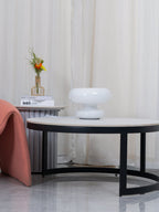 Small unlit Clérachic donut lamp on a distant side table, beyond a round black-framed coffee table and a salmon-pink chair.
