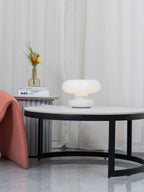 Clérachic donut lamp softly lit on a side table behind a round black coffee table, with a pink chair at the left edge.