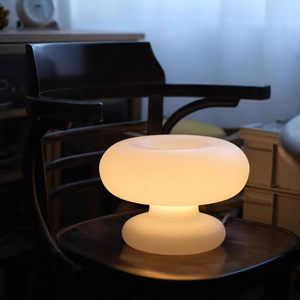 Lit Clérachic donut lamp on a round wooden seat, glowing a warm amber against a dark wooden chair and blue curtains.