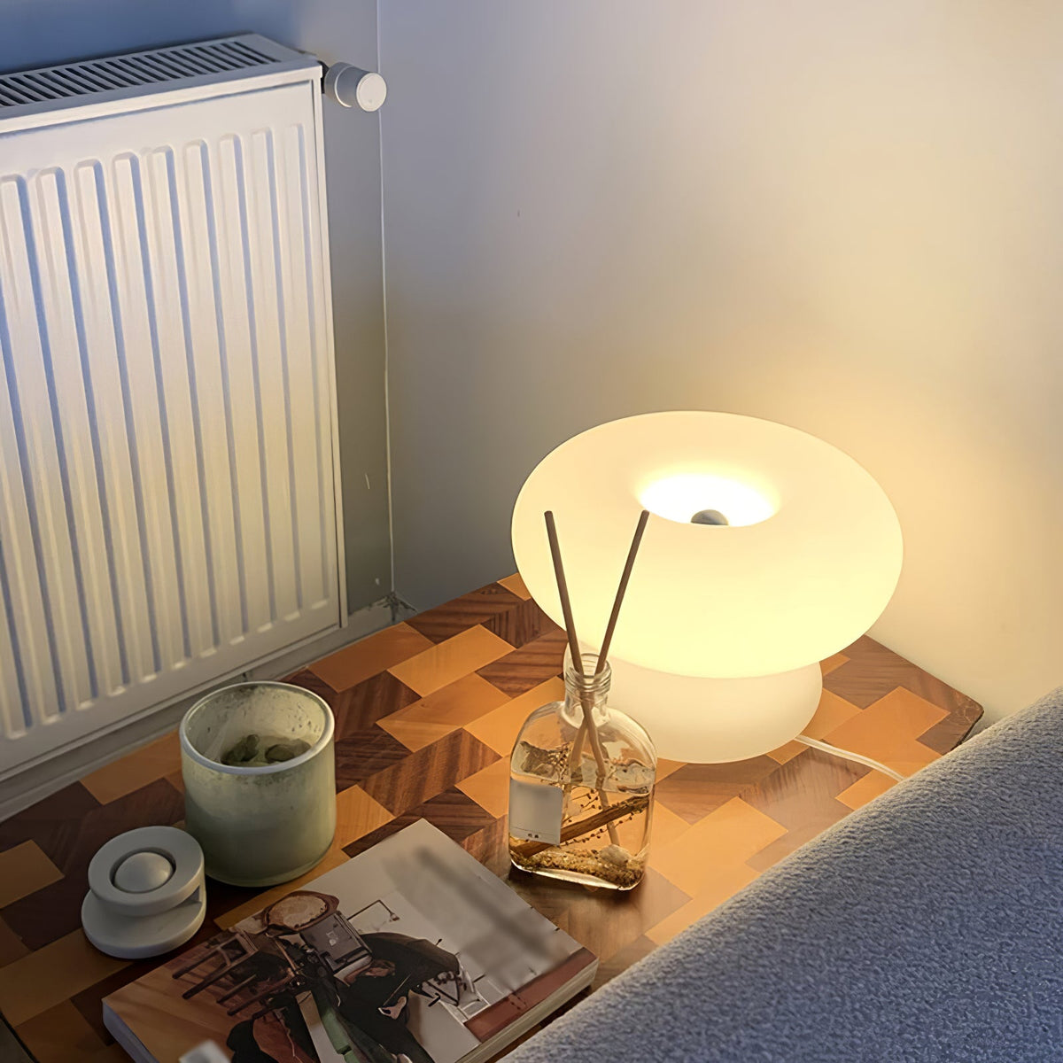 A lit Clérachic donut lamp on a parquet-topped side table, next to a reed diffuser and a candle, with a radiator behind.